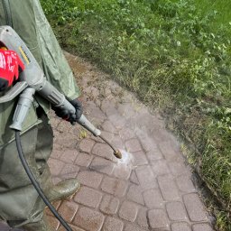 SpecGreen - Czyszczenie kostki brukowej myjką ciśnieniową Kärcher. Osoba w zielonym kombinezonie i gumowcach usuwa zabrudzenia z brązowej kostki, obok widoczna trawa.