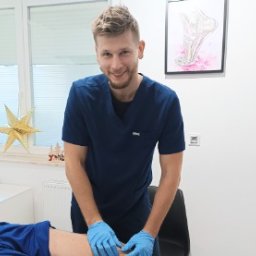 Skowron Med Inowrocław - Fizjoterapia - Rehabilitacja - Masaż - Uśmiechnięty fizjoterapeuta w niebieskim uniformie i rękawicach, wykonuje masaż kolana pacjenta leżącego na stole rehabilitacyjnym w jasnym gabinecie. W tle widoczny plakat z anatomią stawu...