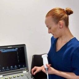 Skowron Med Inowrocław - Fizjoterapia - Rehabilitacja - Masaż - Kobieta w niebieskim uniformie wykonuje badanie USG nadgarstka pacjenta za pomocą ultrasonografu. Widoczny ekran z wynikami badania.