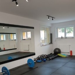 Skowron Med Inowrocław - Fizjoterapia - Rehabilitacja - Masaż - Sala ćwiczeń z dużym lustrem, sztangą z niebieskimi obciążeniami, zestawem kettlebell, piłką do ćwiczeń, matą i grzejnikiem pod oknem.