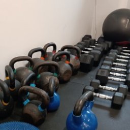 Skowron Med Inowrocław - Fizjoterapia - Rehabilitacja - Masaż - Rząd odważników kettlebell i hantli na czarnej podłodze w siłowni, obok ściany w tle widoczna piłka gimnastyczna.