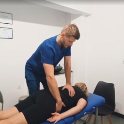 Skowron Med Inowrocław - Fizjoterapia - Rehabilitacja - Masaż - Mężczyzna w niebieskim uniformie wykonuje masaż dolnej części pleców pacjentki leżącej na niebieskim stole do masażu; na ścianie wiszą dwa certyfikaty.