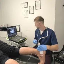 Fizjoterapeuta w niebieskim uniformie wykonuje badanie USG stopy pacjenta leżącego na kozetce, widoczny ekran ultrasonografu i elektrody na skórze.