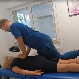 Skowron Med Inowrocław - Fizjoterapia - Rehabilitacja - Masaż - Mężczyzna w niebieskim stroju uciska plecy leżącej na niebieskim łóżku kobiety w czarnym stroju, w tle okno i obrazek stopy.