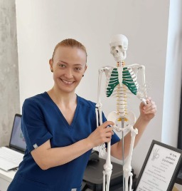 Uśmiechnięta kobieta w niebieskim uniformie prezentuje model szkieletu człowieka w gabinecie w Inowrocławiu. Na biurku widoczny laptop i ulotka informacyjna.