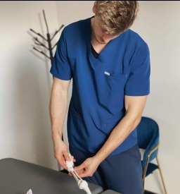 Mężczyzna w niebieskim uniformie demonstruje ruchomość kości stopy na modelu szkieletu, stojąc przy stole w jasnym pomieszczeniu.