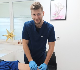 Uśmiechnięty fizjoterapeuta w niebieskim uniformie i rękawicach, wykonuje masaż kolana pacjenta leżącego na stole rehabilitacyjnym w jasnym gabinecie. W tle widoczny plakat z anatomią stawu...