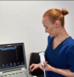 Kobieta w niebieskim uniformie wykonuje badanie USG nadgarstka pacjenta za pomocą ultrasonografu. Widoczny ekran z wynikami badania.