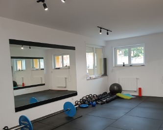 Sala ćwiczeń z dużym lustrem, sztangą z niebieskimi obciążeniami, zestawem kettlebell, piłką do ćwiczeń, matą i grzejnikiem pod oknem.