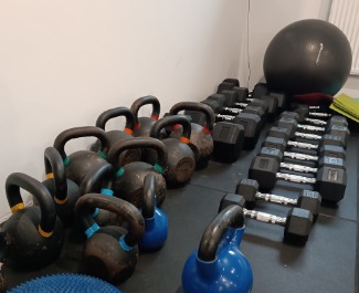 Rząd odważników kettlebell i hantli na czarnej podłodze w siłowni, obok ściany w tle widoczna piłka gimnastyczna.