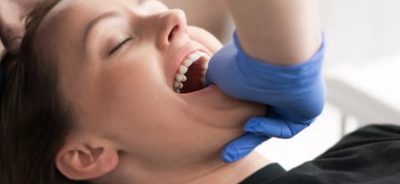 Kobieta z otwartymi ustami podczas terapii manualnej stawu skroniowo-żuchwowego; terapeuta w niebieskich rękawicach uciska szczękę.