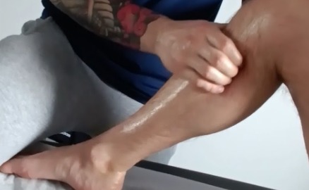 Masażysta dłonią masuje łydkę pacjenta posmarowaną olejkiem, widoczny fragment tatuażu na ramieniu masażysty.