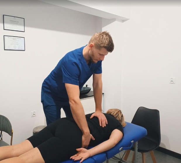Mężczyzna w niebieskim uniformie wykonuje masaż dolnej części pleców pacjentki leżącej na niebieskim stole do masażu; na ścianie wiszą dwa certyfikaty.