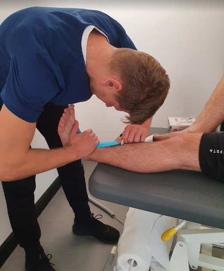 Naklejanie niebieskiej taśmy kinezjologicznej na stopę pacjenta przez terapeutę w gabinecie, widoczny fragment nogi drugiego terapeuty.