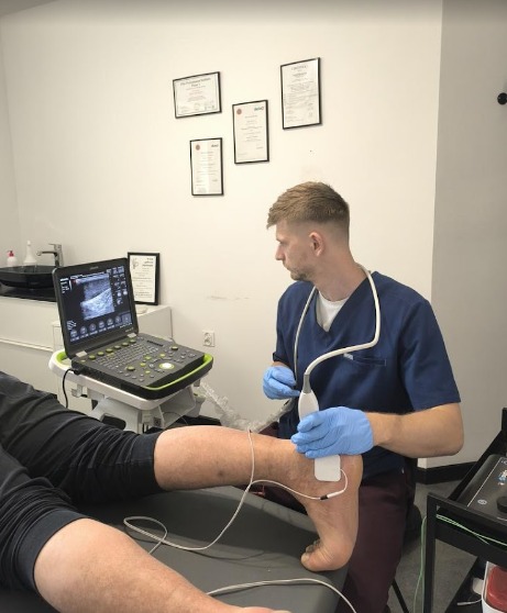 Fizjoterapeuta w niebieskim uniformie wykonuje badanie USG stopy pacjenta leżącego na kozetce, widoczny ekran ultrasonografu i elektrody na skórze.