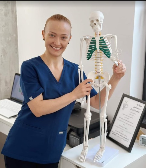 Uśmiechnięta kobieta w niebieskim uniformie prezentuje model ludzkiego szkieletu z zaznaczonymi na zielono żebrami, stojąc obok biurka z laptopem i ramką z tekstem.