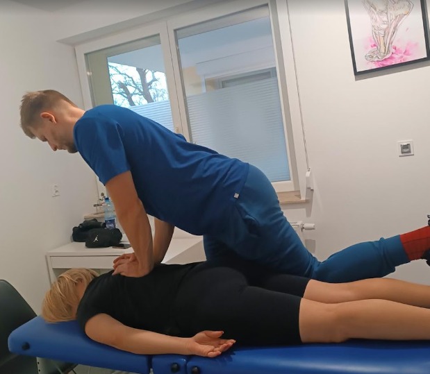 Mężczyzna w niebieskim stroju uciska plecy leżącej na niebieskim łóżku kobiety w czarnym stroju, w tle okno i obrazek stopy.