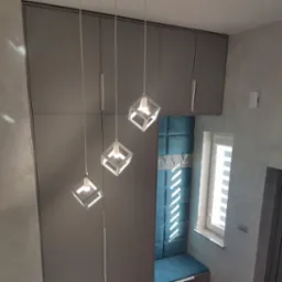 Szara szafa w zabudowie z trzema wiszącymi lampami w kształcie sześcianów, niebieski panel tapicerowany na ścianie z wieszakami, widok z dołu.