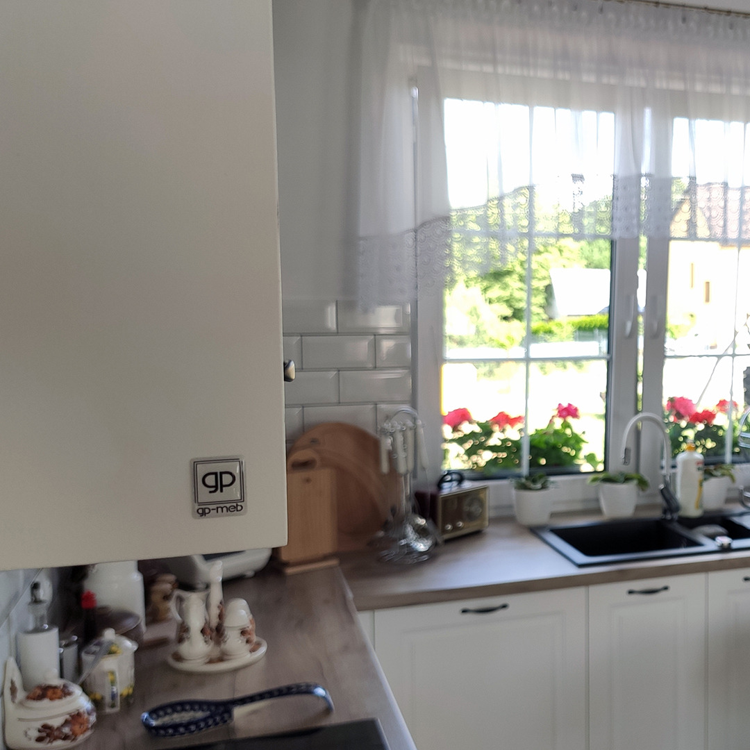 Fragment białej zabudowy kuchennej z widocznym logo producenta, jasny blat, ceramiczne płytki na ścianie i okno z firanką z widokiem na zieleń ogrodu.