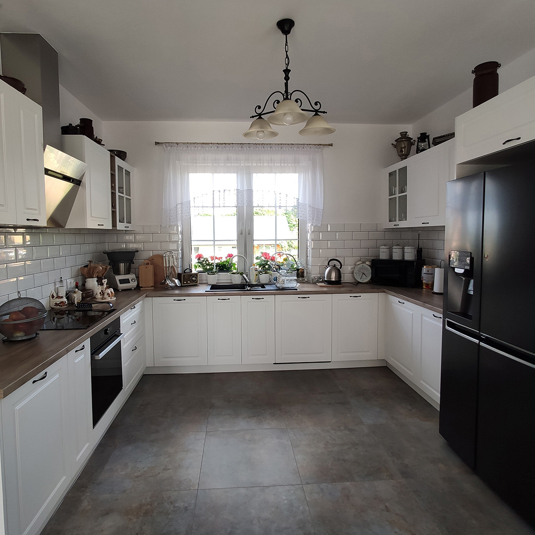 Jasna kuchnia w stylu rustykalnym z białymi frontami szafek, drewnianymi blatami i białymi płytkami na ścianie, okno z firanką i kwiatami w doniczkach, czarna lodówka i ozdobny żyrandol.