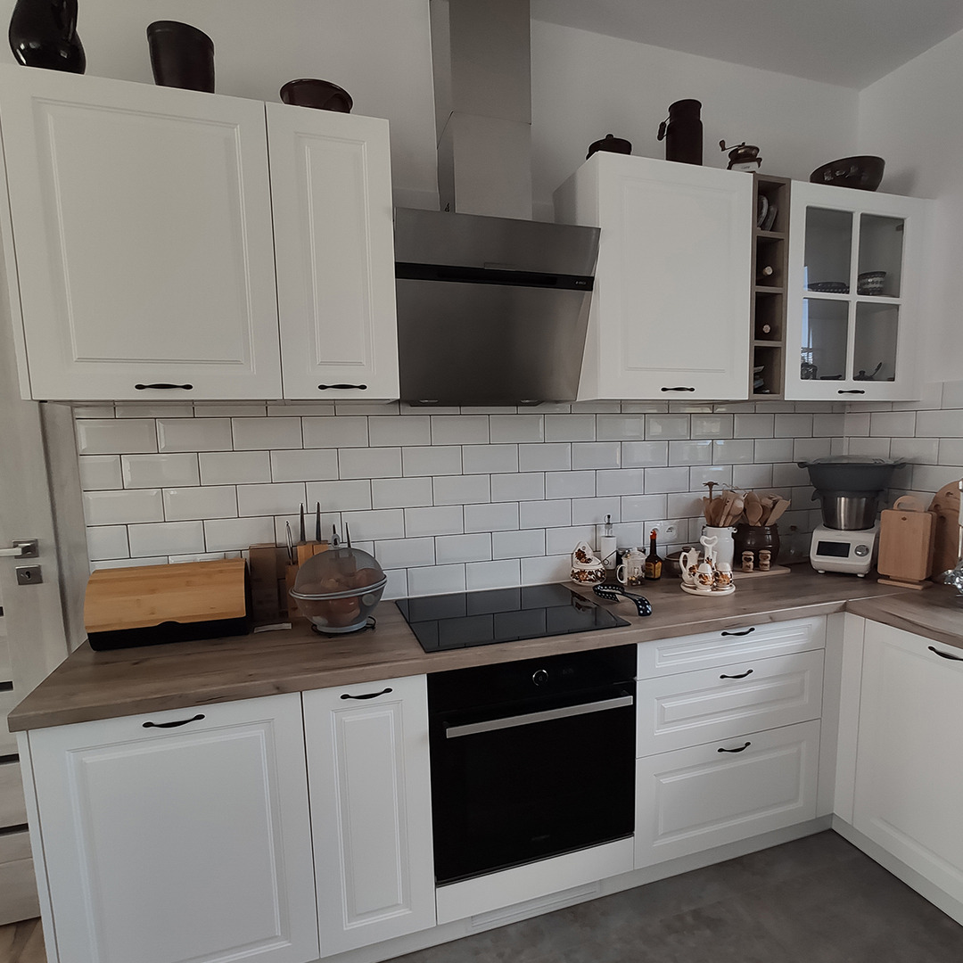 Jasna kuchnia z białymi frontami szafek, czarnymi uchwytami i blatem imitującym drewno. Nad blatem białe płytki ułożone w cegiełkę i srebrny okap.