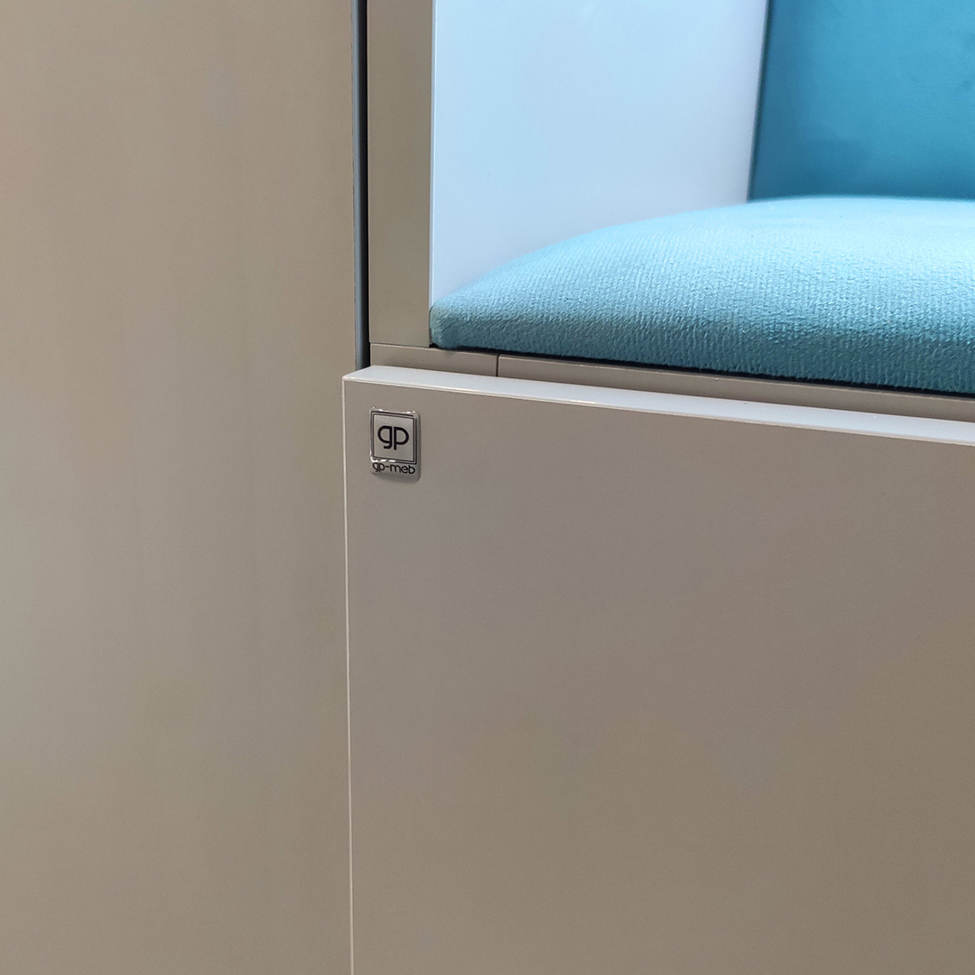 Fragment zabudowy meblowej w odcieniach bieli z siedziskiem wykończonym błękitną tkaniną, widoczne logo producenta na froncie.