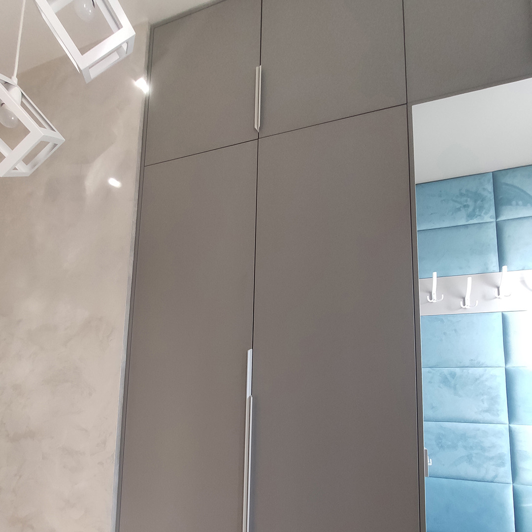 Szara szafa z długimi, srebrnymi uchwytami, widoczna z bliska, z dekoracyjnym oświetleniem i miękkim, niebieskim panelem ściennym w tle.