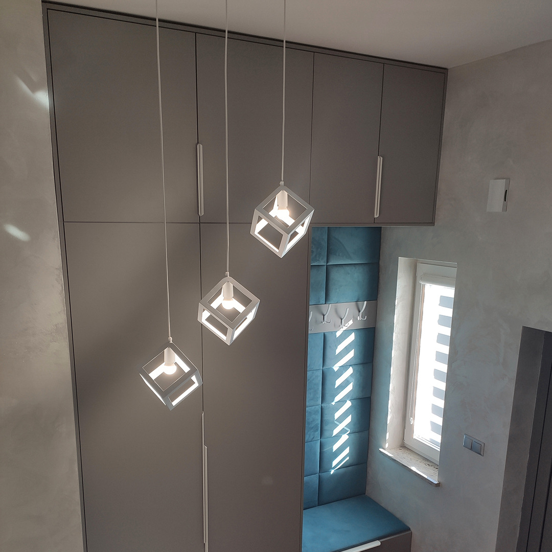 Szara szafa w zabudowie z trzema wiszącymi lampami w kształcie sześcianów, niebieski panel tapicerowany na ścianie z wieszakami, widok z dołu.