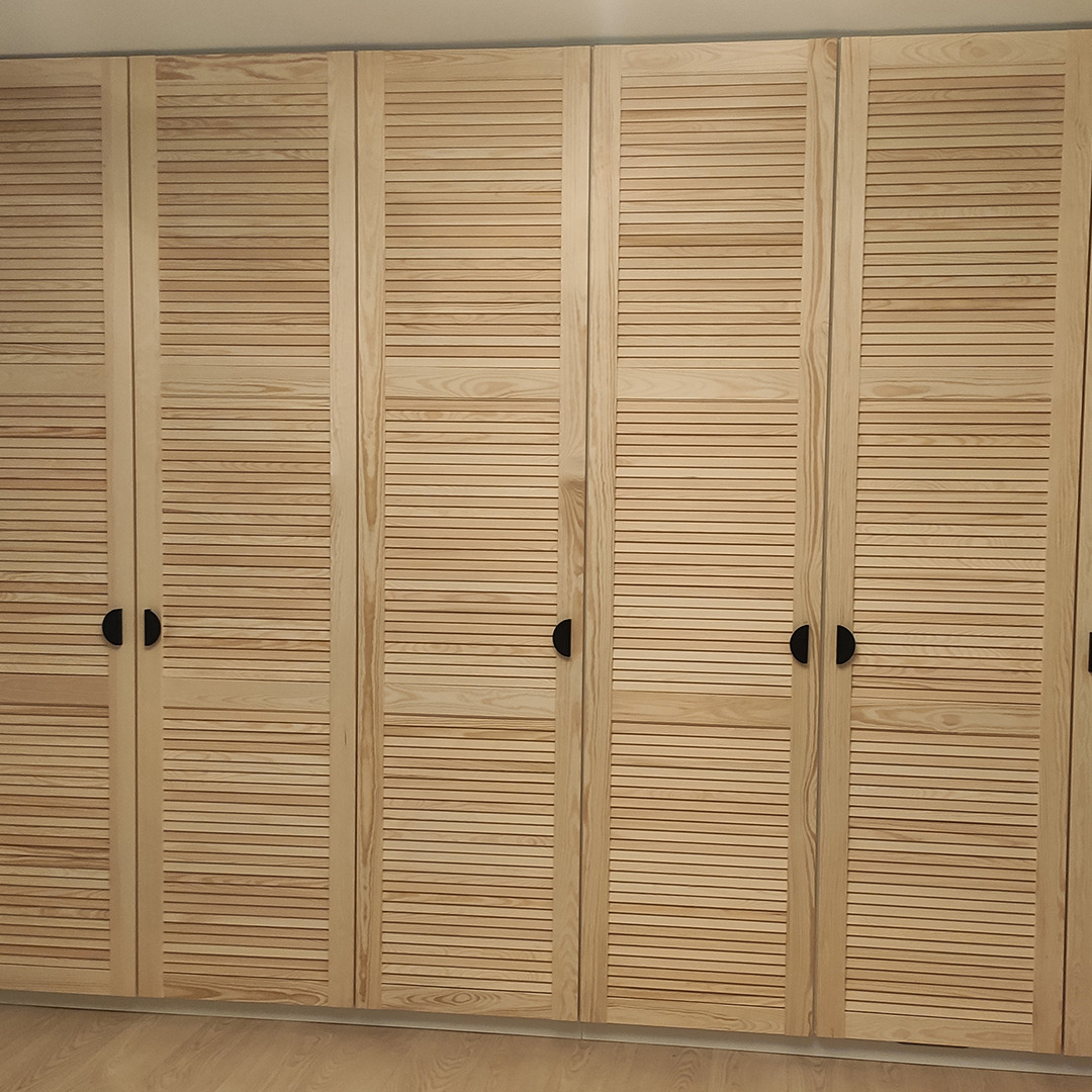 Jasna, drewniana szafa z żaluzjowymi drzwiami i okrągłymi, czarnymi uchwytami, widok z bliska.