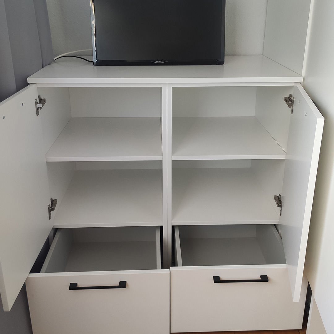 Biała szafka z otwartymi drzwiczkami i szufladami z czarnymi uchwytami, na górze stoi telewizor.