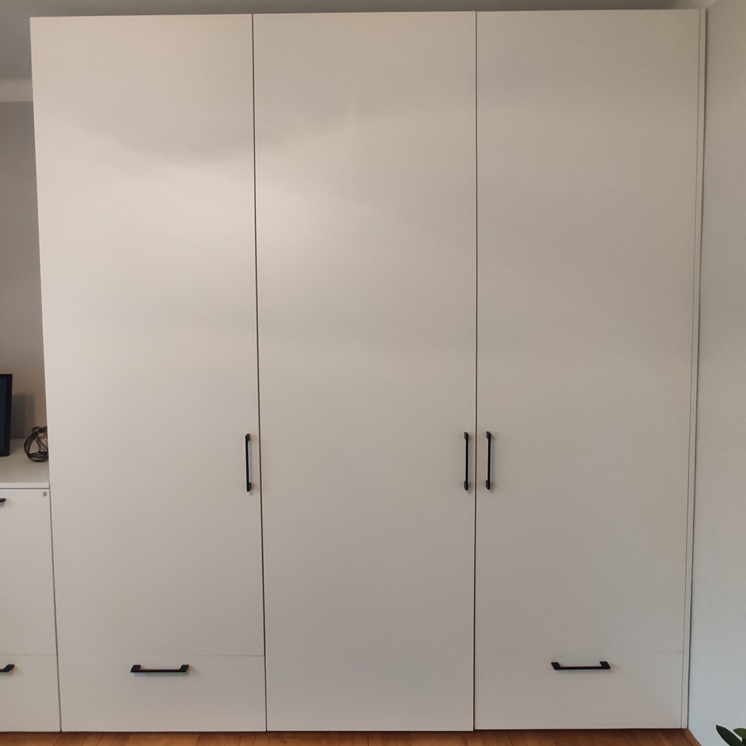 Trzydrzwiowa, matowa szafa wnękowa w kolorze złamanej bieli z podłużnymi, czarnymi uchwytami i szufladami na dole, widoczna na tle jasnej ściany i drewnianej podłogi.