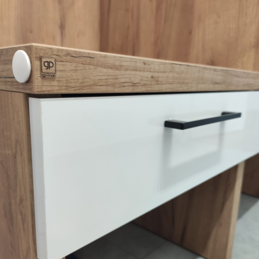 Fragment szafki z białą szufladą i drewnopodobnym blatem z widocznym logo producenta na krawędzi.