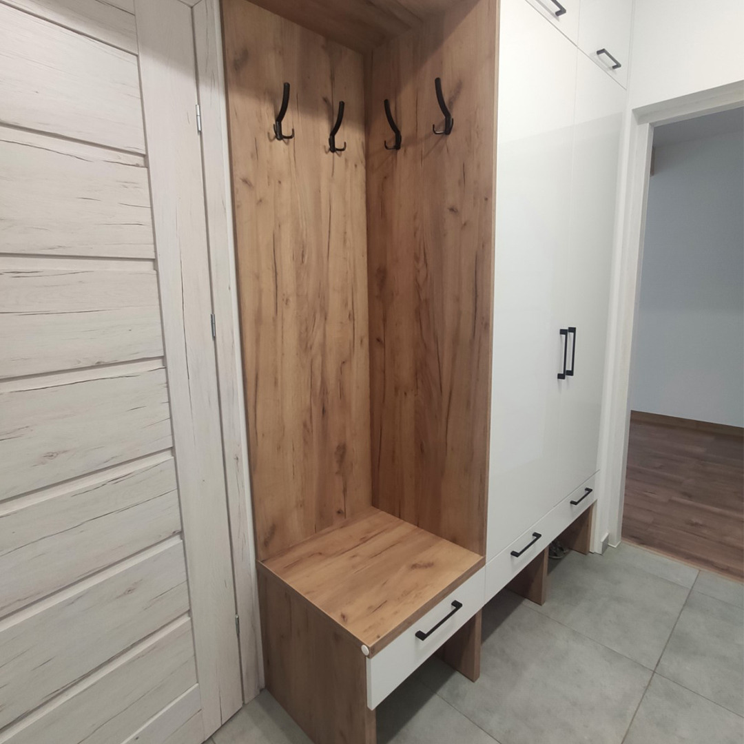 Wnęka w przedpokoju z drewnianym siedziskiem i wieszakami obok białej szafy z czarnymi uchwytami, widok na korytarz w tle.