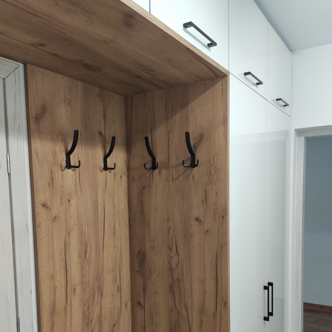 Wnęka z drewnopodobnej płyty z czterema czarnymi haczykami na ubrania, po prawej stronie biała szafa z podłużnymi uchwytami.