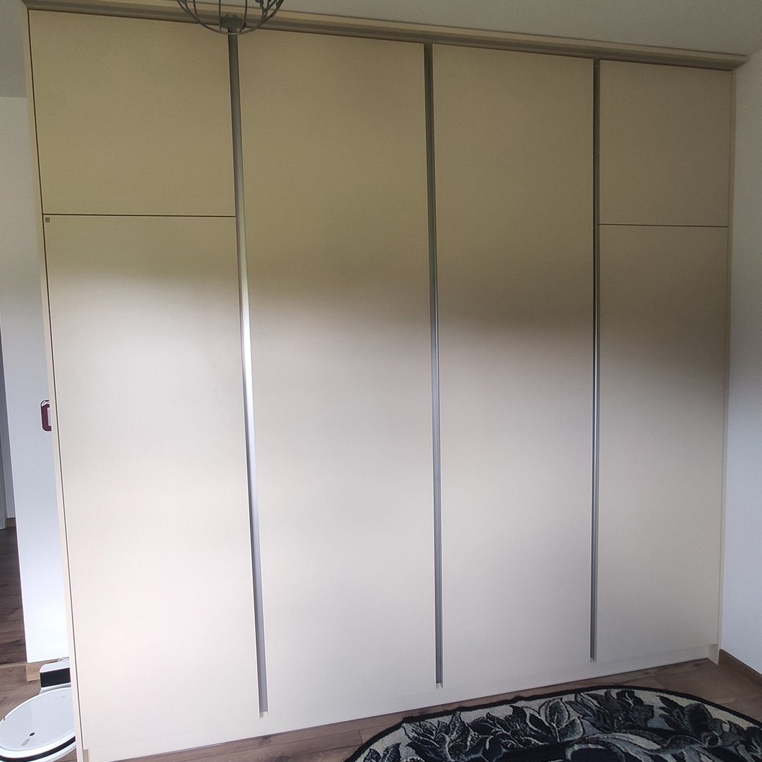 Jasna szafa wnękowa z przesuwnymi drzwiami z metalowymi uchwytami, widoczny robot sprzątający i dywan w tle.