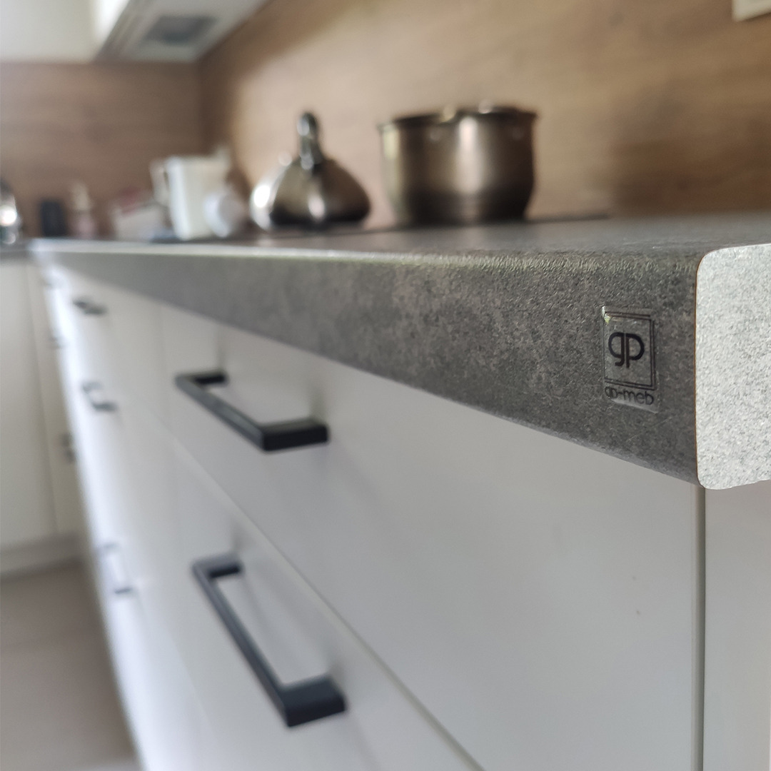 Szary, teksturowany blat kuchenny z zaokrąglonym brzegiem i logo producenta, zamontowany na białych szafkach z czarnymi, podłużnymi uchwytami; w tle widoczne akcesoria kuchenne.