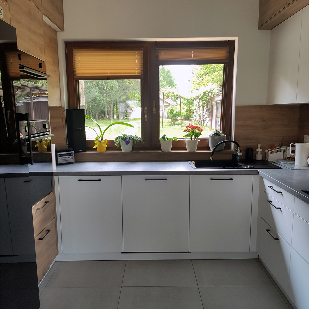 Nowoczesna kuchnia z białymi frontami szafek, szarym blatem i oknem z widokiem na ogród, na parapecie doniczki z kwiatami.