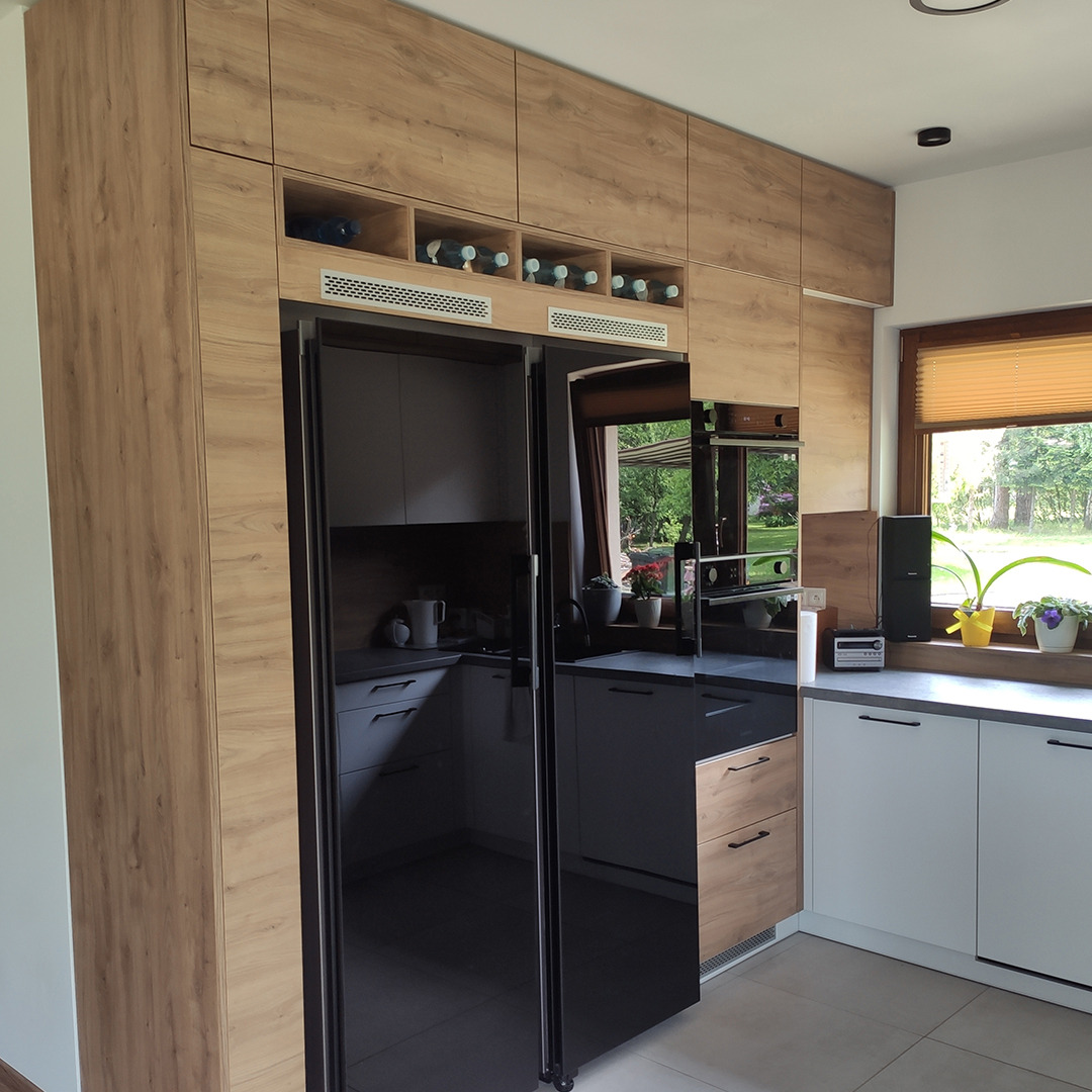 Nowoczesna kuchnia z zabudową drewnopodobną, czarną lodówką i szafkami wiszącymi z półkami na wino, widok na ogród przez okno.