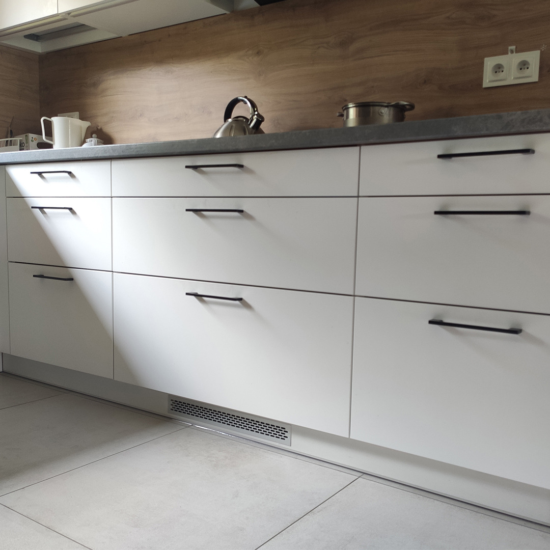 Białe, matowe fronty szuflad kuchennych z czarnymi, minimalistycznymi uchwytami, na blacie czajnik i garnek, drewnopodobna okładzina ściany w tle, widoczna kratka wentylacyjna w cokole.