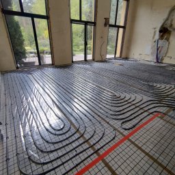 Dominik Człapa - Montaż ogrzewania podłogowego w przestronnym, jasnym pomieszczeniu z dużymi oknami, widoczne rury grzewcze ułożone na siatce, fragment czerwonej taśmy wyznaczającej obszar roboczy, w tle...