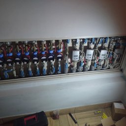 Dominik Człapa - Kolektor rozdzielczy z zaworami, licznikami i przewodami ukryty za klapą rewizyjną. Na podłodze widoczna skrzynka narzędziowa i resztki materiałów.