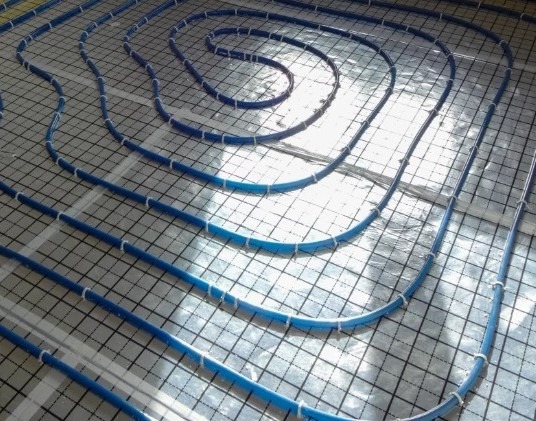 Niebieskie rury ogrzewania podłogowego ułożone na folii z siatką, mocowane białymi opaskami zaciskowymi, widoczne refleksy światła.