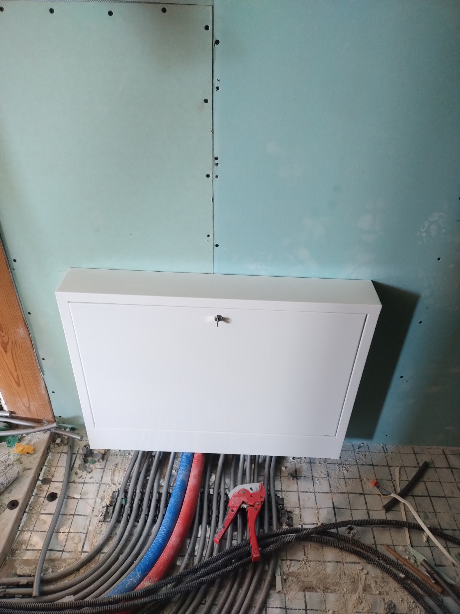 Biała skrzynka rewizyjna na ścianie z płyt kartonowo-gipsowych, pod nią widoczne rury grzewcze w izolacji czerwonej i niebieskiej oraz szare rury instalacji elektrycznej, na podłodze leżą narzędzia...