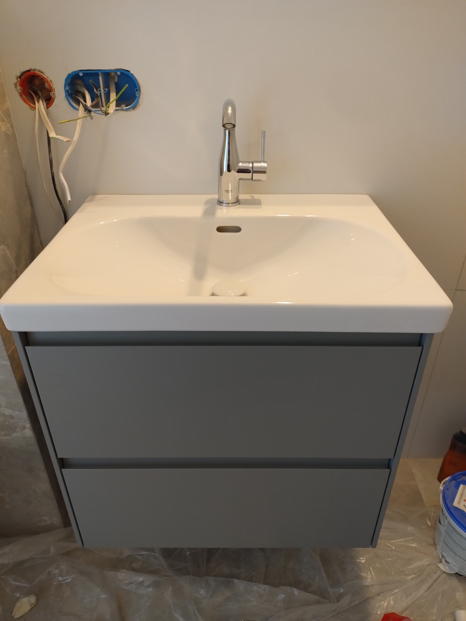 Biała umywalka z szarą szafką podwieszaną i chromowaną baterią, widoczne przewody elektryczne w ścianie, folia ochronna na podłodze.