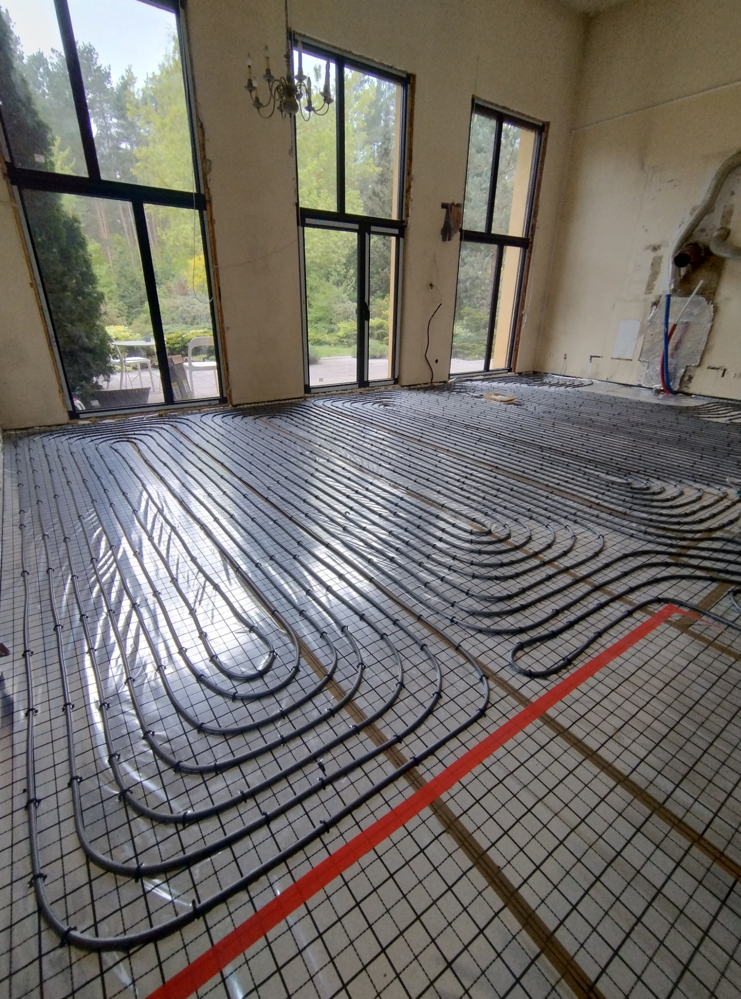 Montaż ogrzewania podłogowego w przestronnym, jasnym pomieszczeniu z dużymi oknami, widoczne rury grzewcze ułożone na siatce, fragment czerwonej taśmy wyznaczającej obszar roboczy, w tle...