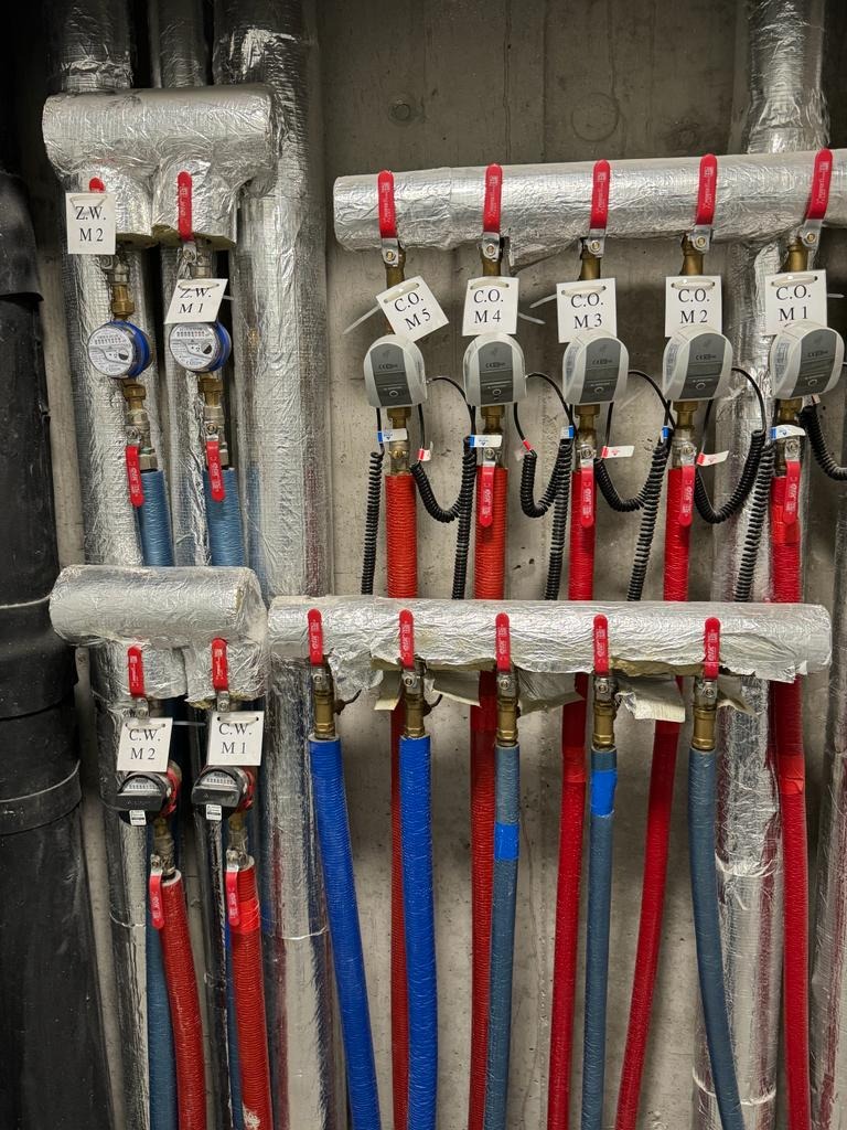 Rozdzielacz instalacji grzewczej z rurami w kolorach czerwonym i niebieskim, izolowanymi termicznie, z zamontowanymi licznikami ciepła oznaczonymi jako C.O. M1-M5 oraz Z.W. M1-M2 i C.W. M1-M2,...