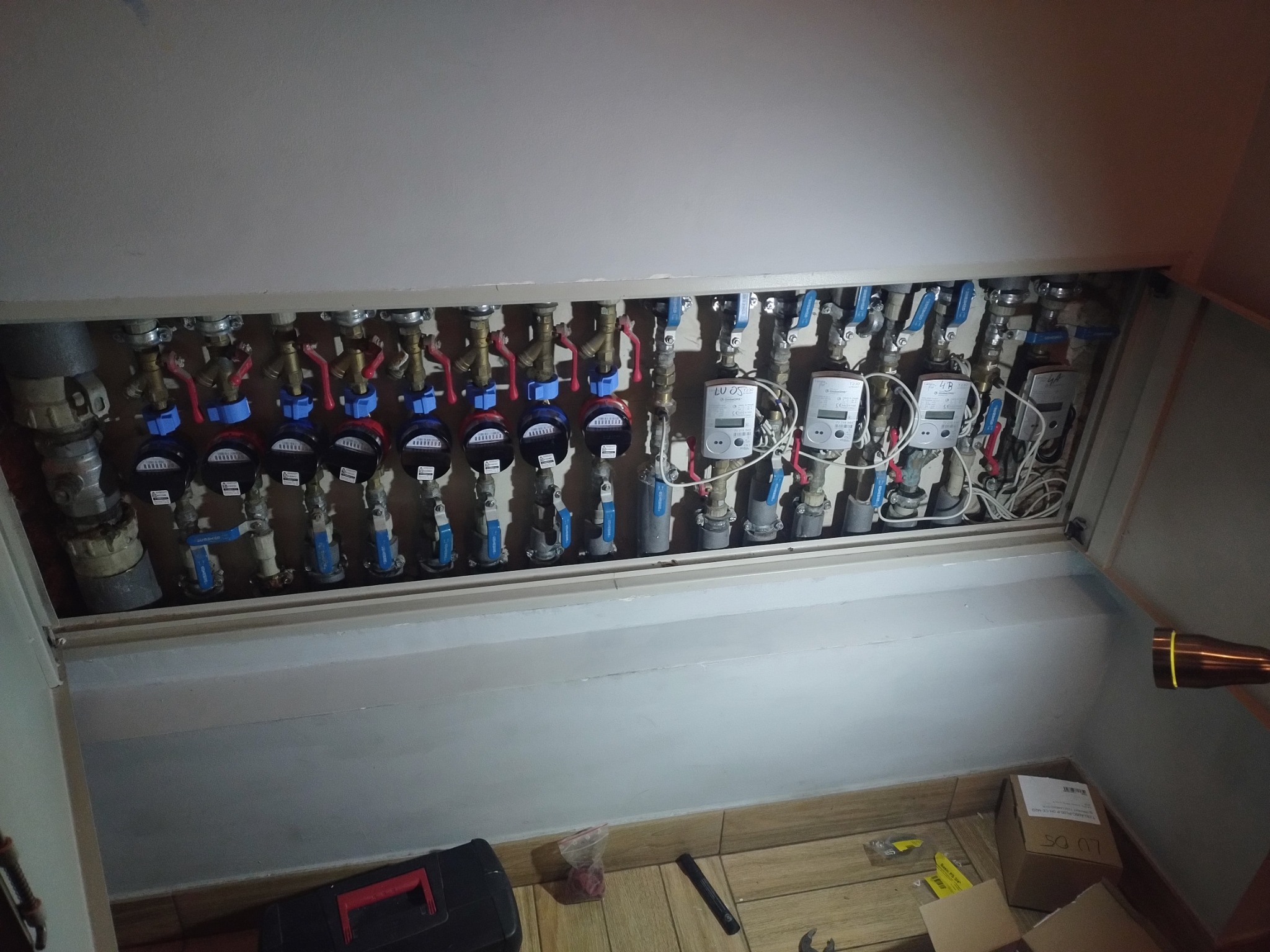 Kolektor rozdzielczy z zaworami, licznikami i przewodami ukryty za klapą rewizyjną. Na podłodze widoczna skrzynka narzędziowa i resztki materiałów.