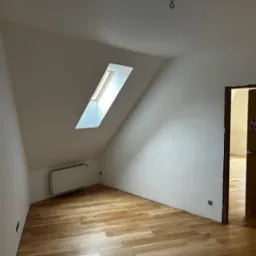 Jasne poddasze z oknem dachowym, świeżo pomalowane ściany na biało, drewniana podłoga. Widoczny grzejnik i fragment drzwi z dekoracjami.