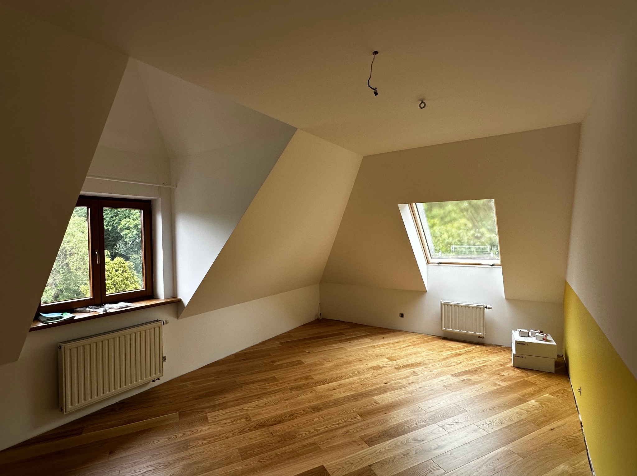 Jasne poddasze po remoncie: drewniana podłoga, białe ściany, okna dachowe i grzejniki. Fragment żółtej ściany i kartony w rogu dodają charakteru.