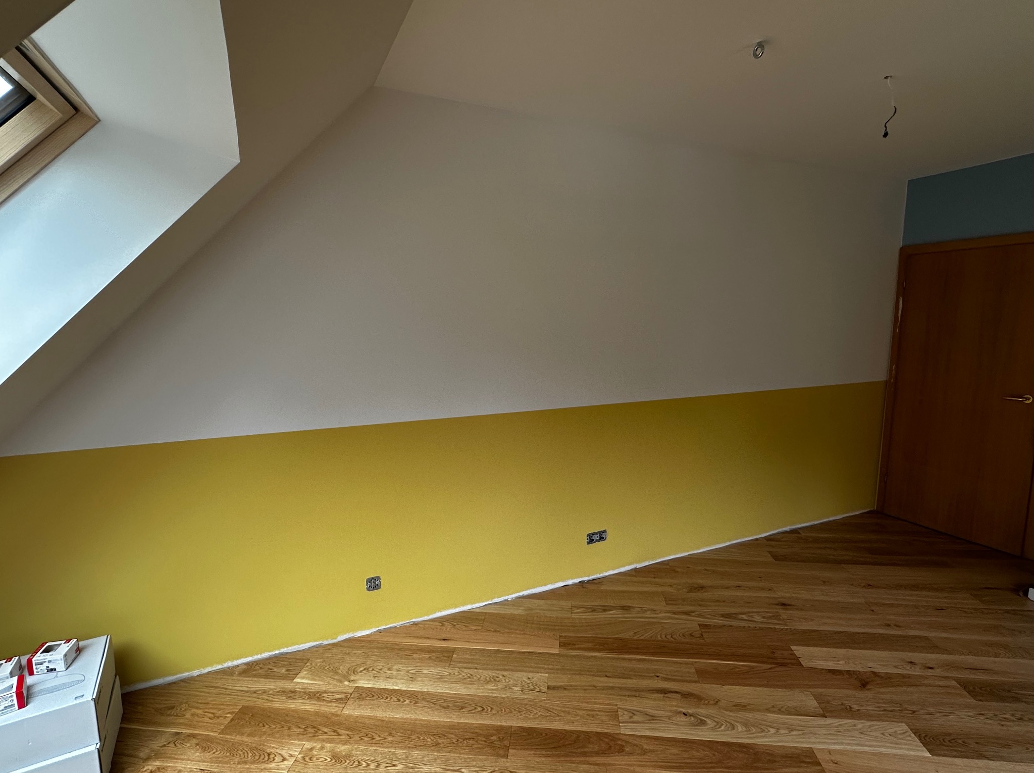 Pomieszczenie po malowaniu: żółta dolna część ściany, biała górna, skośny sufit z oknem dachowym, drewniana podłoga. Widoczne puszki elektryczne.